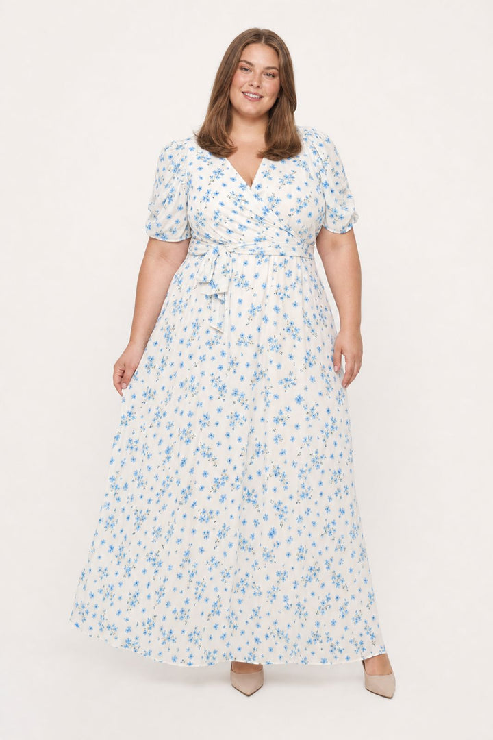 Plus-size kvinde i en lang hvid maxikjole med blåt blomsterprint mod en hvid baggrund.