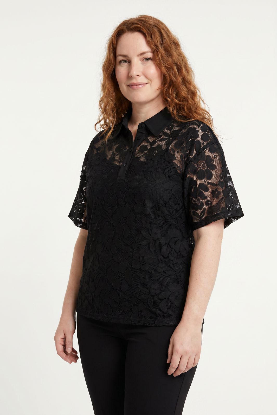 Rødhåret kvinde iført en sort poloshirt i blomstret blonder mod en hvid baggrund.