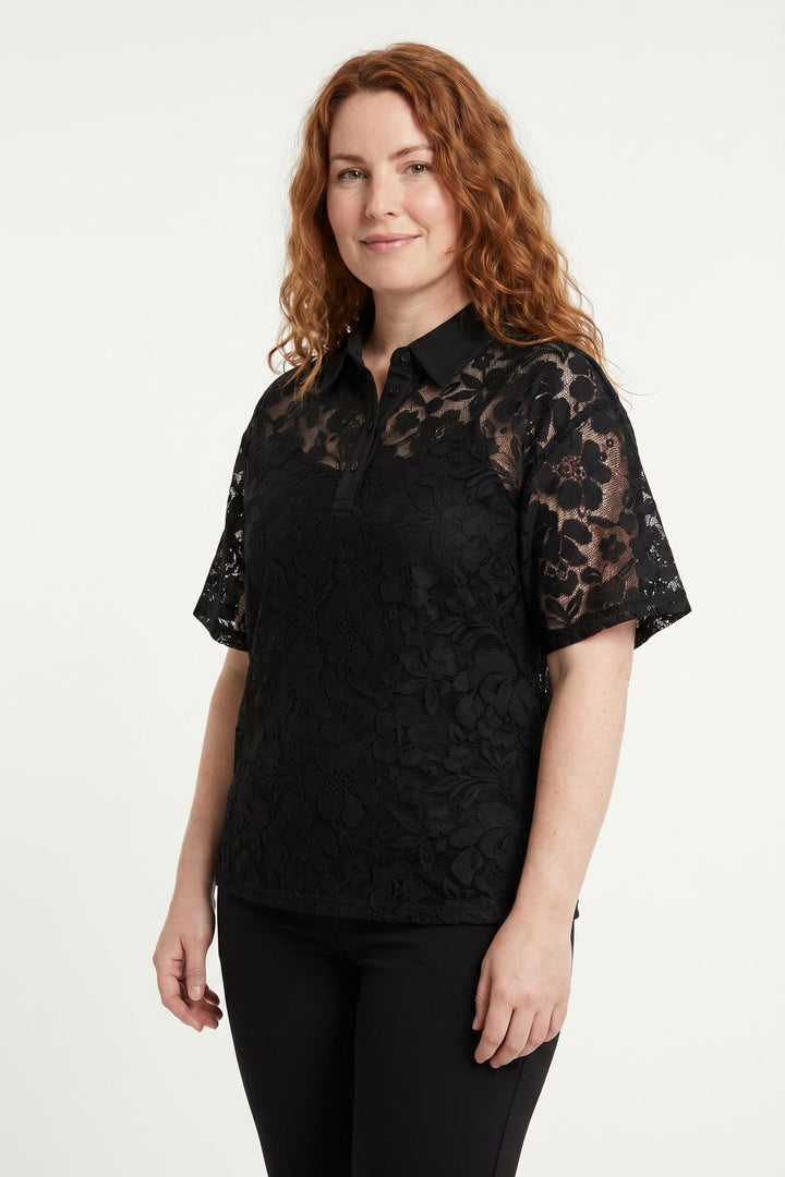 Rødhåret kvinde iført en sort poloshirt i blomstret blonder mod en hvid baggrund.