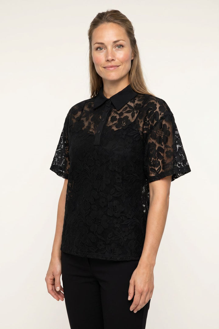 Kvinde iført en sort kortærmet blonde-poloshirt med blomstermønster mod en hvid baggrund.