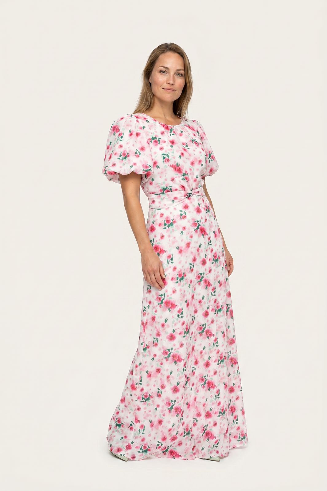 Kvinde i en lang hvid kjole med lyserødt blomsterprint og pufærmer mod en lys baggrund.