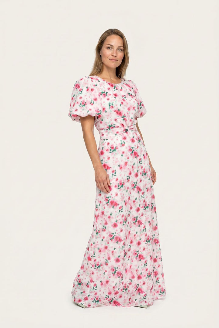 Kvinde i en lang hvid kjole med lyserødt blomsterprint og pufærmer mod en lys baggrund.