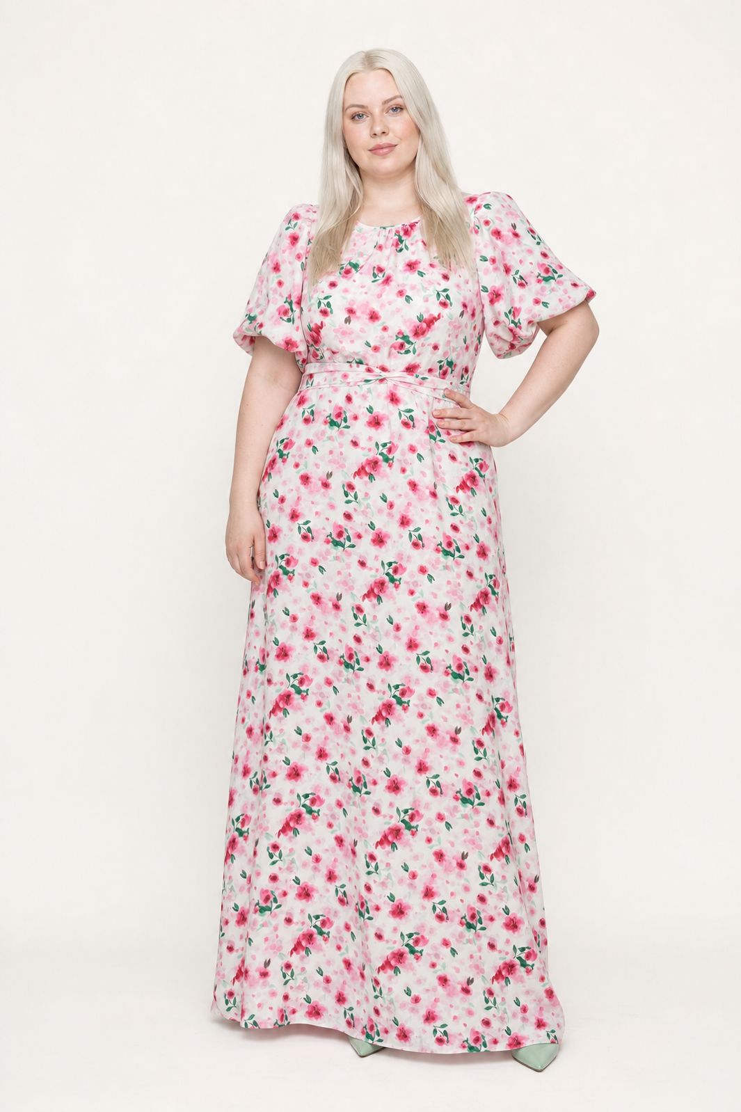 Blond kvinde i en lang hvid kjole med lyserødt blomsterprint og pufærmer mod en hvid baggrund.
