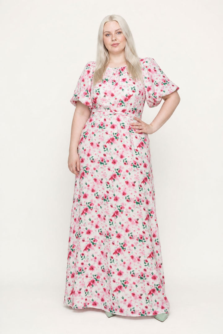 Blond kvinde i en lang hvid kjole med lyserødt blomsterprint og pufærmer mod en hvid baggrund.